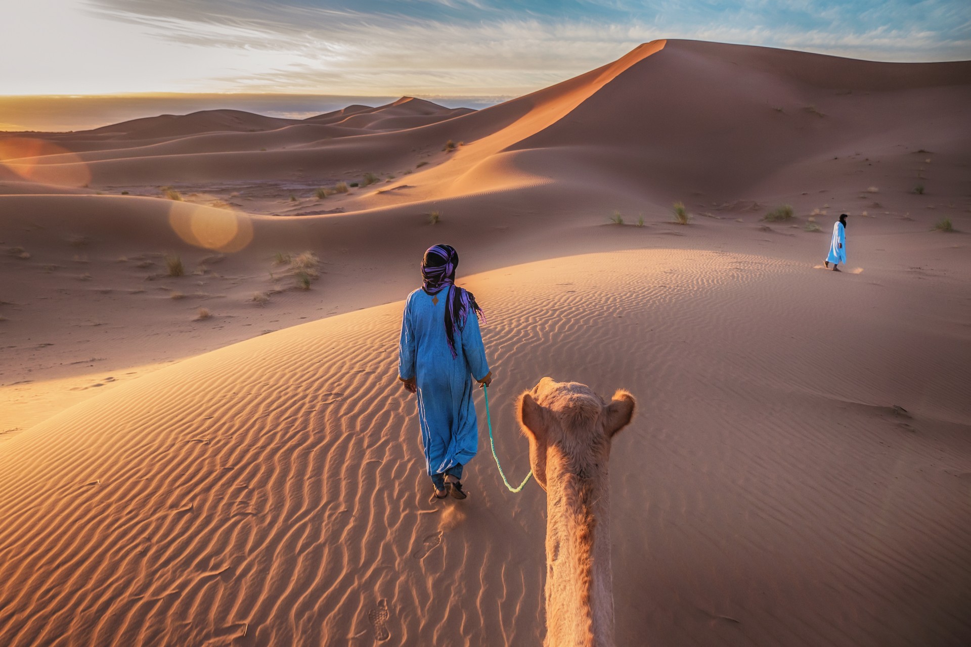 Showing the traditional blue clothing and mode of transportation of nomadic Tuareg tribesmen in the Sahara Desert, as they walk through the sand dunes with camels.