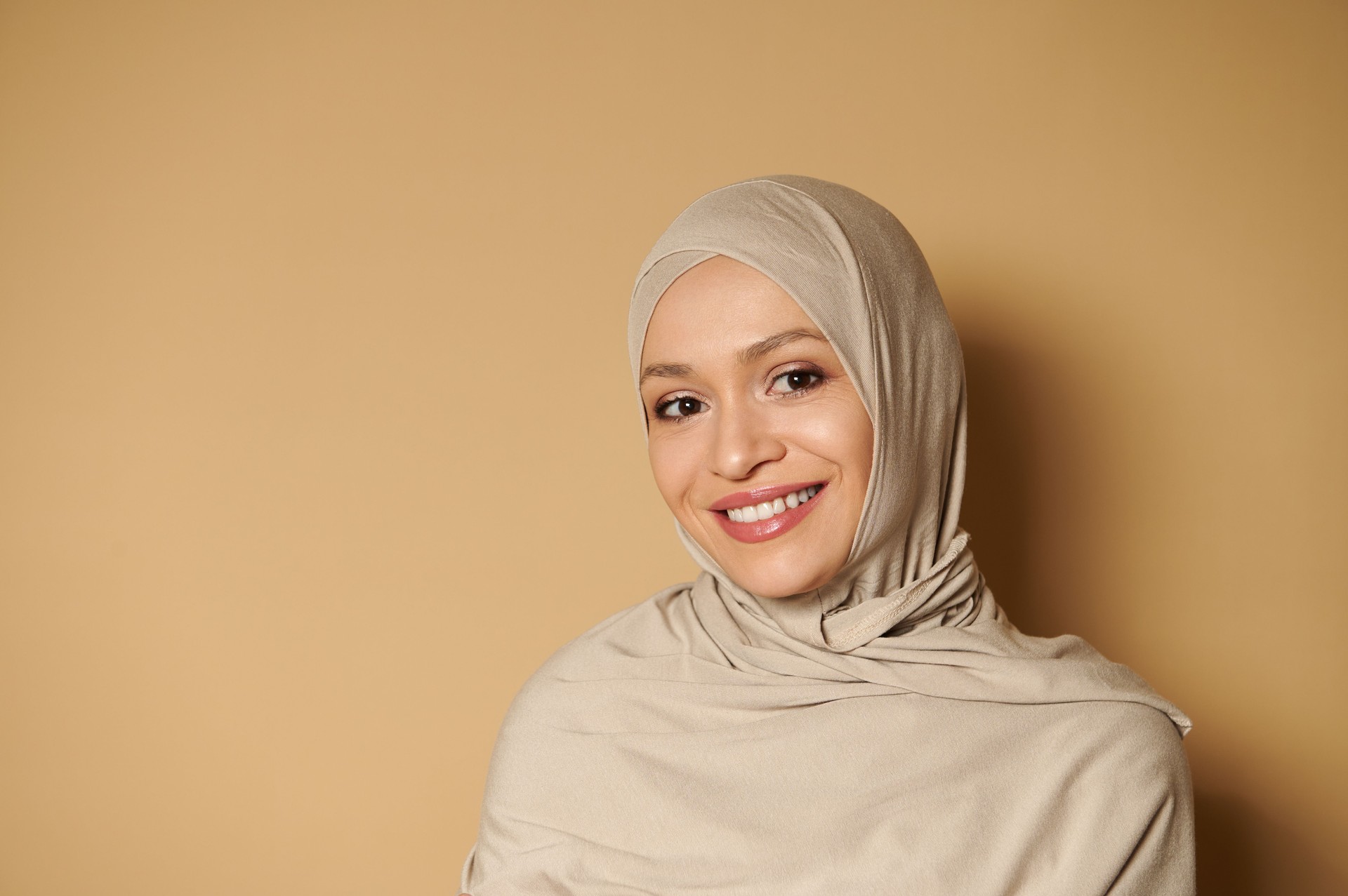 Arab muslim woman wearing hijab looking at camera while posing with a beautiful toothy smile on a beige background with copy space.