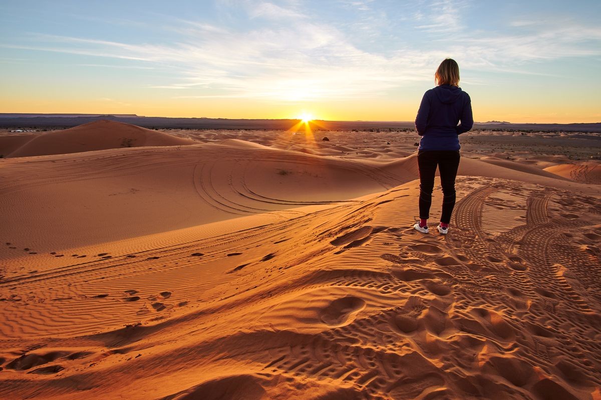 Blonde tourist girl watching sunrise in Sahara desert on top of the sand dune in Merzouga Morocco Africa