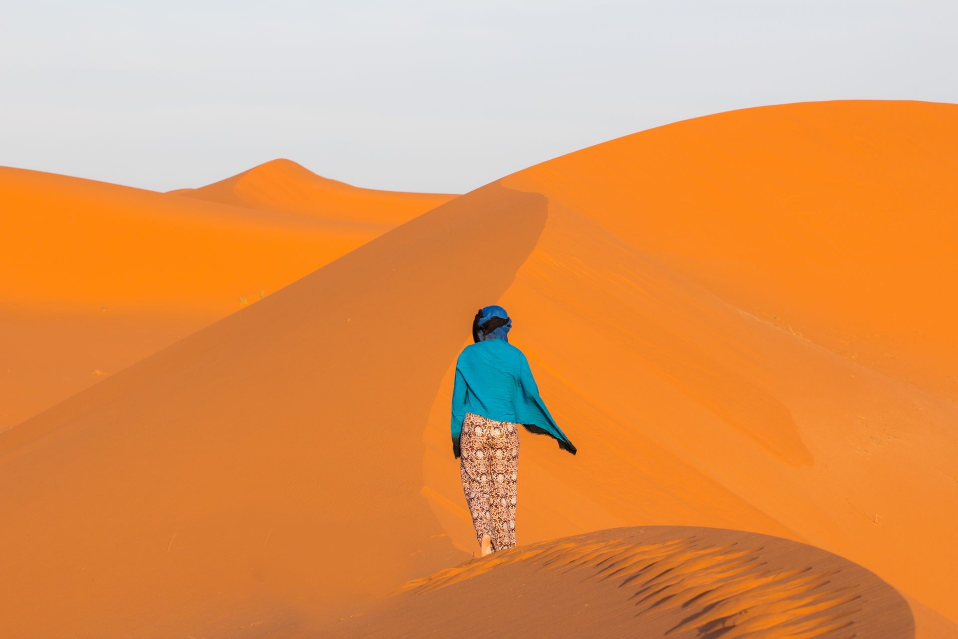 Girl in blue scarf is going on the deserts dune back to the camera and looking at yellow sands of Sahara in Morocco